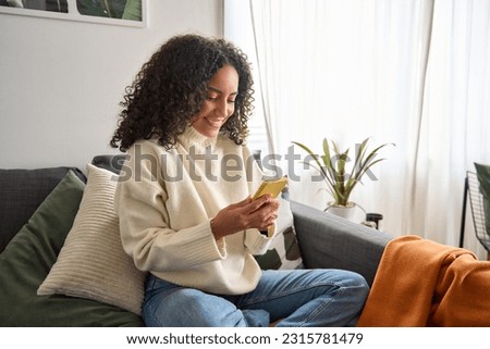 Happy young latin woman sitting on sofa holding mobile phone using cellphone technology doing ecommerce shopping, buying online, texting messages relaxing on couch in cozy living room at home. Royalty-Free Stock Photo #2315781479