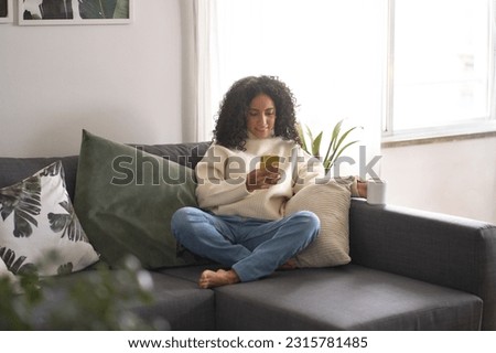 Relaxed young latin woman sitting on sofa using cell phone at home holding smartphone, looking at cellphone doing shopping in mobile digital apps buying ecommerce products online on couch. Royalty-Free Stock Photo #2315781485