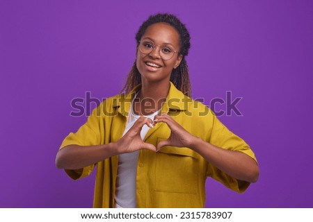 Young beautiful romantic African American woman makes heart shaped gesture to confess her love to boyfriend on eve of valentines day holiday stands on purple background. Relationship, intimacy