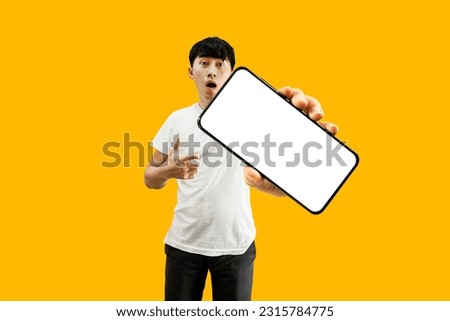 Asian Man Hand Pointing At Empty White Smartphone Screen on yellow Background. Cellphone Display Mock Up For Mobile App Advertisement.