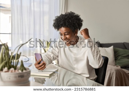Excited African American teen student feeling excited winner holding cell using mobile phone winning online, receiving good news on smartphone getting reward celebrating scholarship at home. Royalty-Free Stock Photo #2315786881