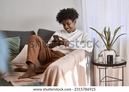 Happy gen z African American teen sitting on couch holding smartphone, using cell phone, looking at cellphone enjoying doing online ecommerce shopping in mobile apps, chatting or playing games.
