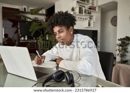 African American student elearning online class writing notes at home. Teenage boy using computer watching webinar, learning virtual lesson, taking hybrid web course writing notes sitting at table.