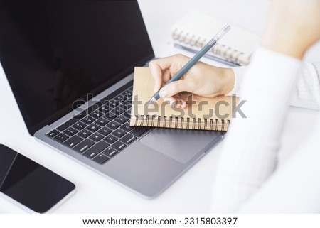 Close-up capture of a woman's hand as she writes in a notepad on a sleek laptop, with an out-of-focus background