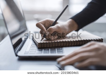 Detailed view of a woman's hand taking notes in a notepad on a cutting-edge laptop, against a blurred backdrop