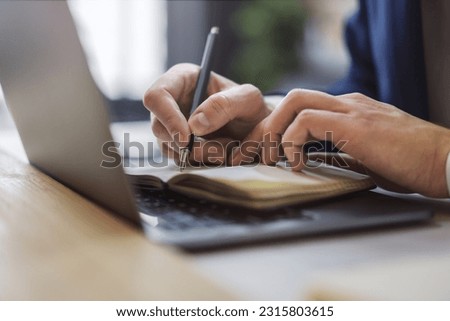 Detailed view of a man's hand jotting down notes in a notepad on a modern laptop, with a blurred background