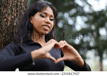 A black woman stands near a tree, showing a heart with her hands.