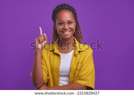 Young cheerful resourceful African American woman pointing finger up with sincere smile as she came up with idea of launching own startup to achieve financial success stands on purple background Royalty-Free Stock Photo #2315828427