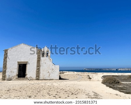 The picture shows a small whitewashed church standing on the cliffs of the Algarve near Sagres. The sky is bright blue and in the background you can see the ocean and the cliffs of the coast. 