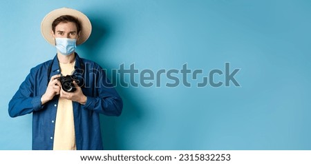 Covid-19, pandemic and travel concept. Happy guy on vacation taking pictures with camera, wearing medical mask and summer hat, blue background.