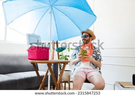 Happy man with hat and sunglasses drinking a pineapple drink while enjoying the summer at home pretending to be at the beach Royalty-Free Stock Photo #2315847253