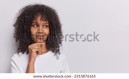 Studio shot of pretty woman with dark curly hair keeps hand under chin daydreams about something thinks about pleasant has tricky plan dressed in casual t shirt isolated on white background copy space Royalty-Free Stock Photo #2315876165