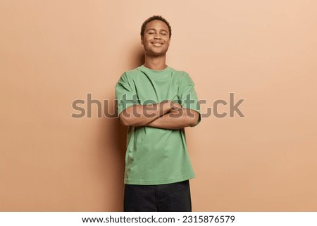 Isolated studio shot of dark skinned young man stands self confident keeps arms folded dressed in casual green t shirt and black trousers feels proud of himself poses against brown background. Royalty-Free Stock Photo #2315876579