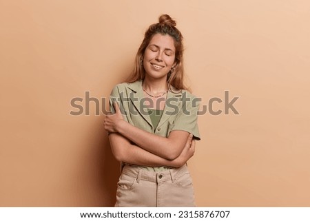 European girl wears shirt trousers and embraces herself with warm smile sstands isolated against beige background emanating sense of inner peace and self acceptance isolated over brown background Royalty-Free Stock Photo #2315876707