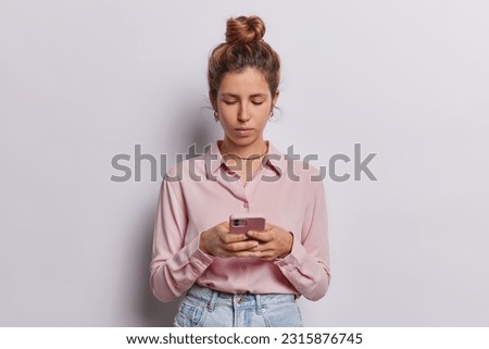 Horizontal shot of serious attentive European woman with piercing in nose uses mobile phone for chatting online sends text messages wears formal shirt and jeans isolated over white background.
