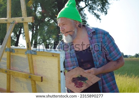 Midsection trendy enthusiastic painter working outdoors having creative inspiration painting picture holding paintbrush in mouth. Mature man artist hipster creating artwork oil painting at meadow dawn