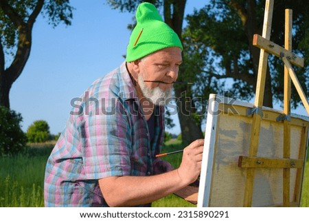 Midsection trendy enthusiastic painter working outdoors having creative inspiration painting picture holding paintbrush in mouth. Mature man artist hipster creating artwork oil painting at meadow dawn