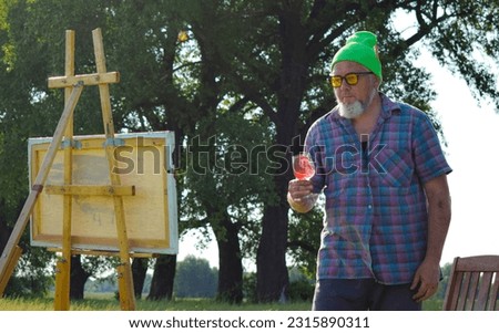 Creative male person contemplates oil painting on wooden easel drinks wine celebrates finalizing of his work outdoors. Good wine helps to relax painter, take a pause in art process at meadow sun light