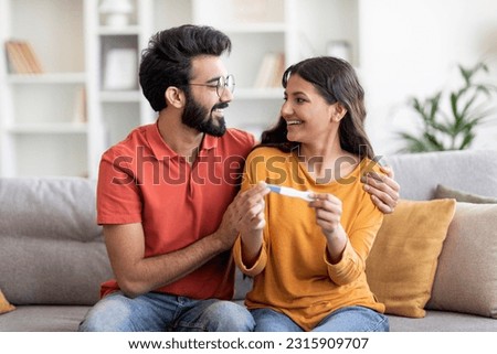 Happy indian couple holding positive pregnancy test and embracing at home, romantic young eastern spouses waiting for baby, celebrating becoming parents, sitting on couch in living room, closeup
