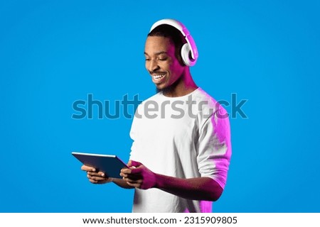 Entertainment and modern technologies. Happy attractive young african american guy in white shirt using modern digital tablet and wireless headphones in neon light over blue background, copy space Royalty-Free Stock Photo #2315909805
