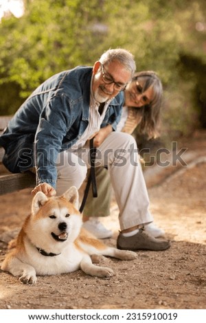 Positive european mature couple sit on bench with dog, enjoy date together, walk, petting animal in park at weekend in summer, outdoor, vertical. Pet love, free time, training Royalty-Free Stock Photo #2315910019