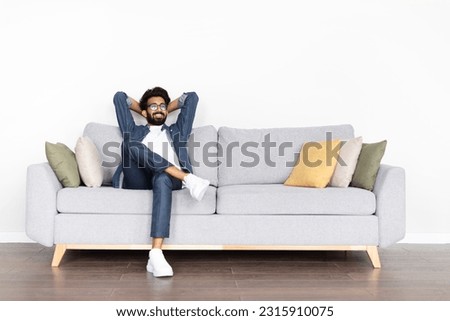 Relaxed happy young eastern guy wearing eyeglasses and casual comfy outfit chilling alone on couch at home, looking at copy space and smiling, enjoying his new house, resting at weekend Royalty-Free Stock Photo #2315910075