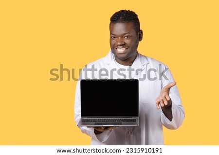 Telemedicine. Toothy black doctor man in white coat uniform showing laptop computer with empty screen, recommending medical website, mockup, standing over yellow studio background
