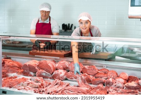 Smiling skilled young female butcher working behind counter in butchery, showing fresh raw veal loin slices Royalty-Free Stock Photo #2315955419