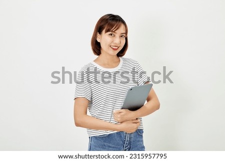 Charming young Asian female employee with short brown hair wearing t-shirt and jeans smiling and holding digital tablet standing on white background copy space