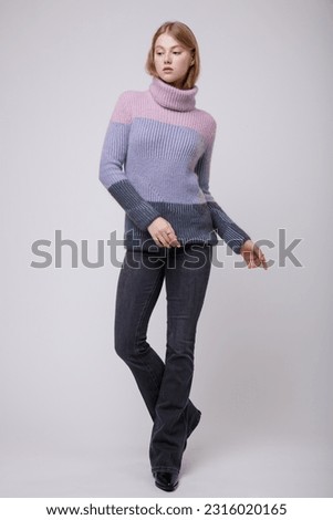 Fashion photo of beautiful elegant young woman in a pretty triple color pink, blue, gray sweater, blouse, black jeans, denim posing on white, soft gray background. Blonde
