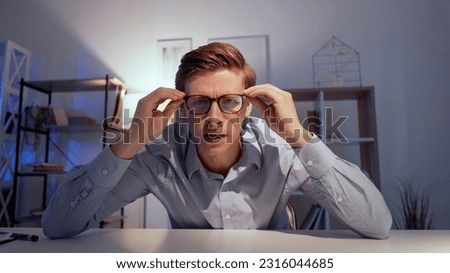 Shocked man. Online conference. Interesting news. Smart curious guy sitting work desk looking on camera holding glasses in light room interior. Royalty-Free Stock Photo #2316044685