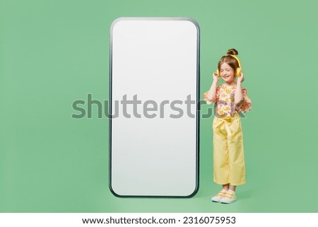 Full body little child kid girl 6-7 years old wear casual clothes big blank screen area mobile cell phone listen music in headphones isolated on plain green background Mother's Day love family concept
