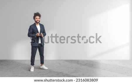 Full length portrait of cheerful young Middle Eastern businessman in elegant suit holding smartphone standing in empty room. Concept of business and communication. Copy space