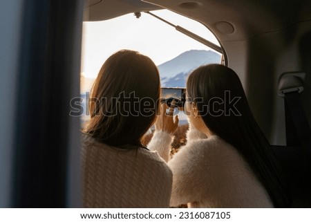 Happy Asian woman friends enjoy and fun outdoor activity lifestyle using mobile phone taking picture of Mt Fuji covered in snow at autumn sunset during travel nature road trip on car at Kawaguchi lake