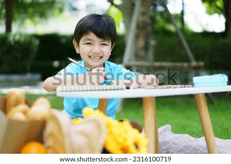 Happy cheerful Asian little boy enjoy drawing and painting alone at home backyard alone, boy drawing a picture at outdoor garden portrait on a beautiful natural bokeh background.