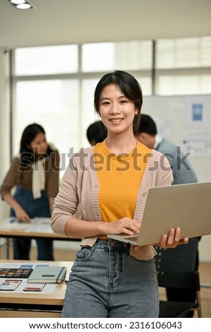 A portrait of a beautiful and charming young Asian female graphic designer or website developer stands in the co-working office with a laptop in her hand.