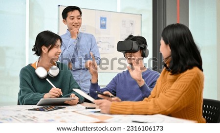 A cheerful and excited young Asian male developer sitting at the table tested a new VR game with virtual reality goggles in the meeting with his team. Business tech startup company concept