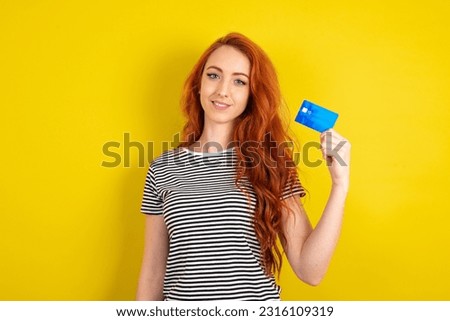 Photo of happy cheerful smiling positive young beautiful red haired woman wearing striped shirt over yellow studio background recommend credit card