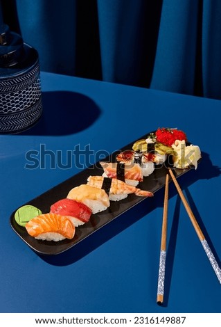Japanese sushi - A set of sushi with salmon, tuna, shrimp, eel, and avocado on a black plate on a deep blue background with chopsticks. Bright light, harsh shadows. A modern still life, minimalist