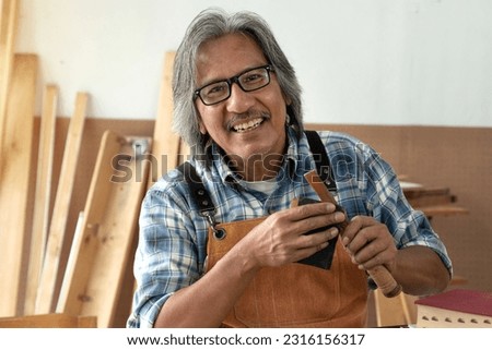 Happy senior carpenter smiling at camera and holding carpentry tool in hand, business owner of wood work shop