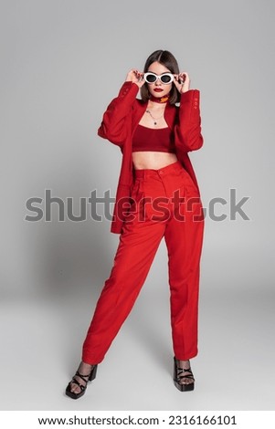 generation z, tattooed young woman with short hair and nose piercing posing in sunglasses and red suit on grey background, modern fashion, trendy outfit, chic style, full length Royalty-Free Stock Photo #2316166101