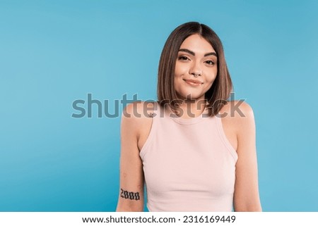 not knowing, smiling young woman with tattoos and nose piercing standing in tank top on blue background, looking at camera, confused, pretty face, generation z, summer outfit Royalty-Free Stock Photo #2316169449