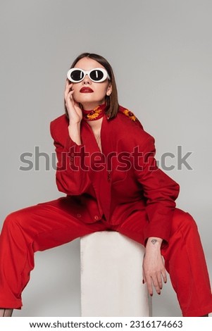 generation z, fashion model with brunette short hair and nose piercing posing in sunglasses and red suit while sitting on concrete cube on grey background, lady in red, young woman in red outfit Royalty-Free Stock Photo #2316174663