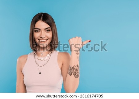 cheerfulness, tattooed young woman with short brunette hair in tank top smiling and pointing with thumb on blue background, casual attire, gen z fashion, happiness, showing something on camera Royalty-Free Stock Photo #2316174705