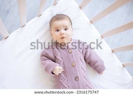 a newborn baby in a crib in a brown warm jumpsuit on a white cotton bed has woken up in the morning or is falling asleep, a surprised portrait of a baby Royalty-Free Stock Photo #2316175747