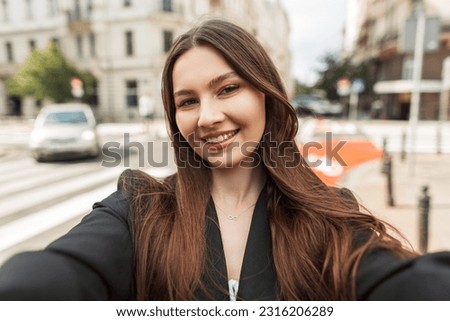 Happy beautiful young chic lady with a smile in fashion clothes with a black blazer walks in the city and takes a selfie on the camera