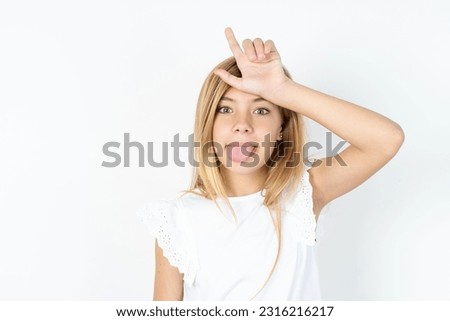 beautiful caucasian teen girl wearing white T-shirt over white wall gestures with finger on forehead makes loser gesture makes fun of people shows tongue