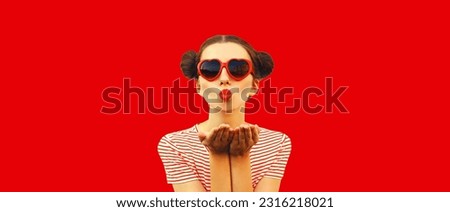 Portrait of stylish young woman blowing her lips with lipstick sending sweet air kiss wearing red heart shaped sunglasses with cool girly hairstyle on background