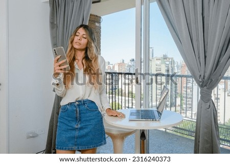 young latina woman of colombian ethnicity, in the living room at home reading messages from her clients on the phone, standing next to the laptop leaning on the table. business concept, copy space.