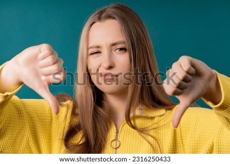Portrait of unhappy european woman condemns with sign of dislike. Young millennial lady expressing discontent with showing thumbs-down gesture on blue studio background. Royalty-Free Stock Photo #2316250433